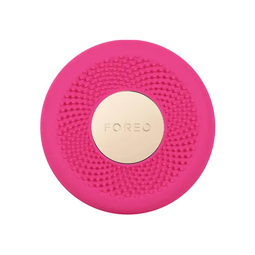 A circular pink facial cleansing brush head with a textured silicone scrubber surface and a central gold-toned metallic button embossed with the brand name FOREO.