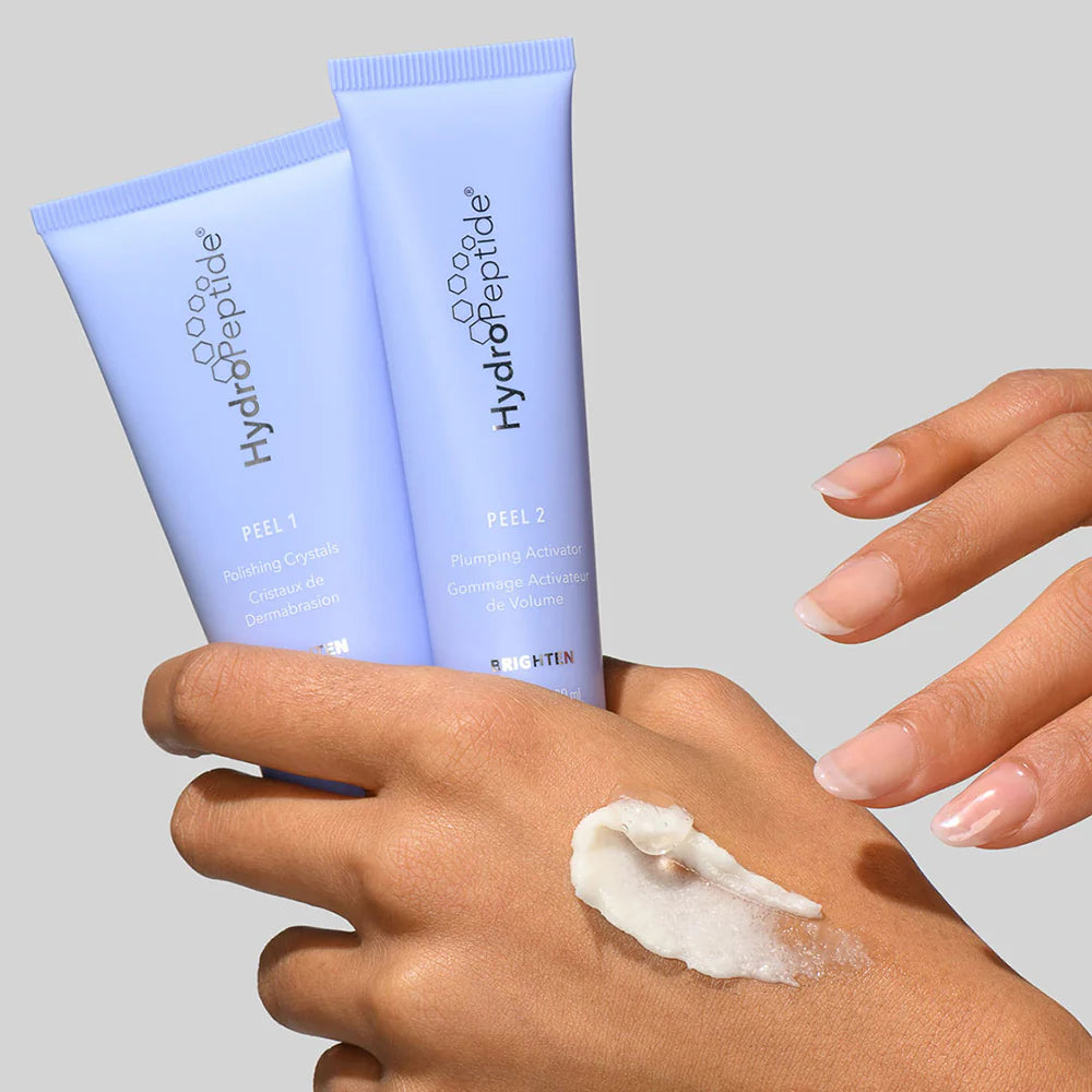 Two tubes of HydroPeptide skincare product on a gray background with a hand applying cream.