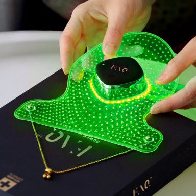 A person's hands are shown holding a glowing green LED light therapy panel over a black box containing a gold necklace. The panel is illuminated, casting a yellow ring of light, and the brand name 'FAO' is visible on both the central black device and the box.