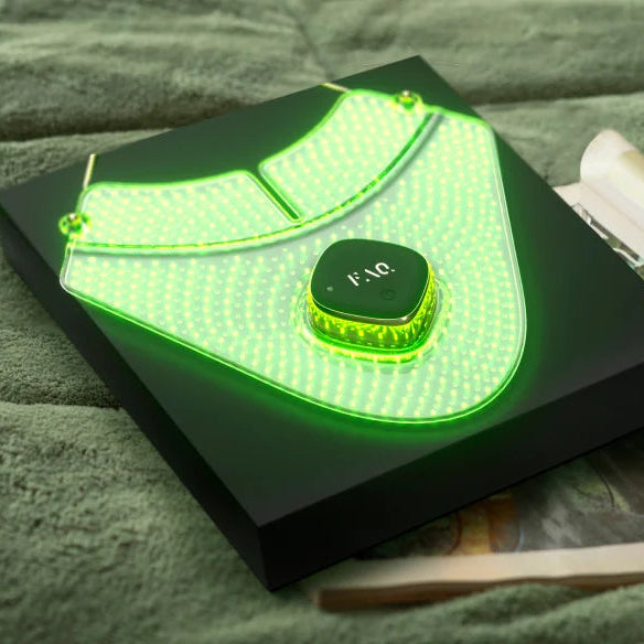 A black rectangular electronic device with a glowing green LED neck light accessory attached, resting on a textured green fabric surface next to an open magazine. The device has a central circular control labeled 'FAB'.