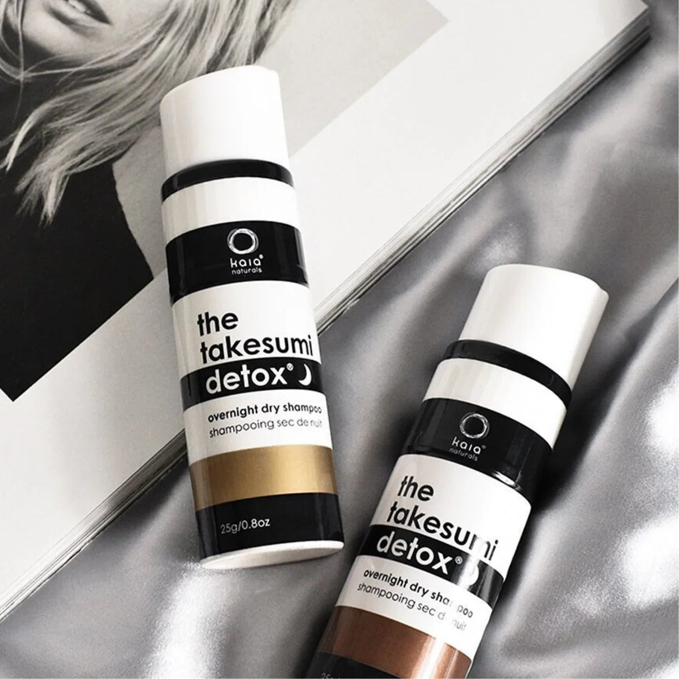 Two bottles of 'the takesumii detox' dry shampoo on a reflective surface with a magazine in the background.