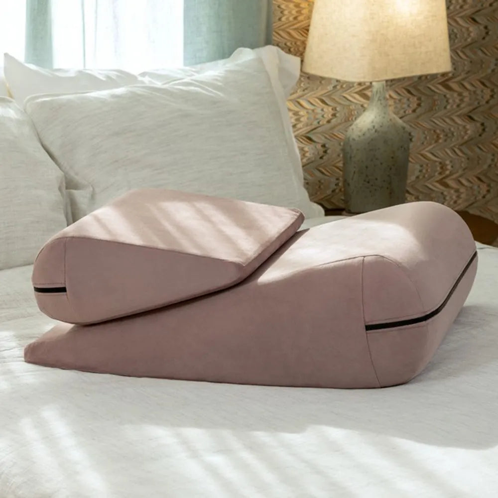 Pink wedge pillow on a bed with a lamp and curtains in the background