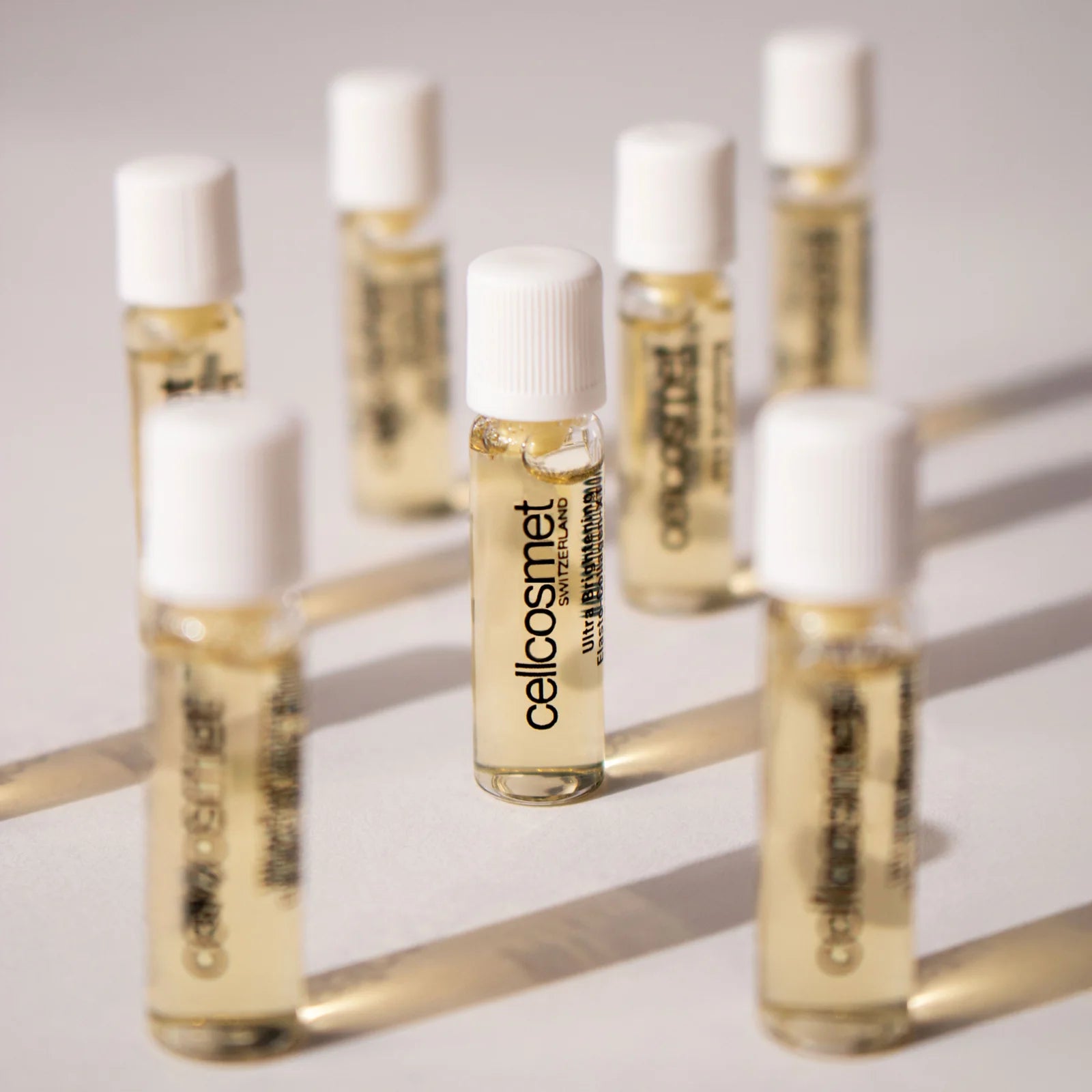 Set of small Cellcosmet glass bottles with white caps, containing a clear serum, on a light background.