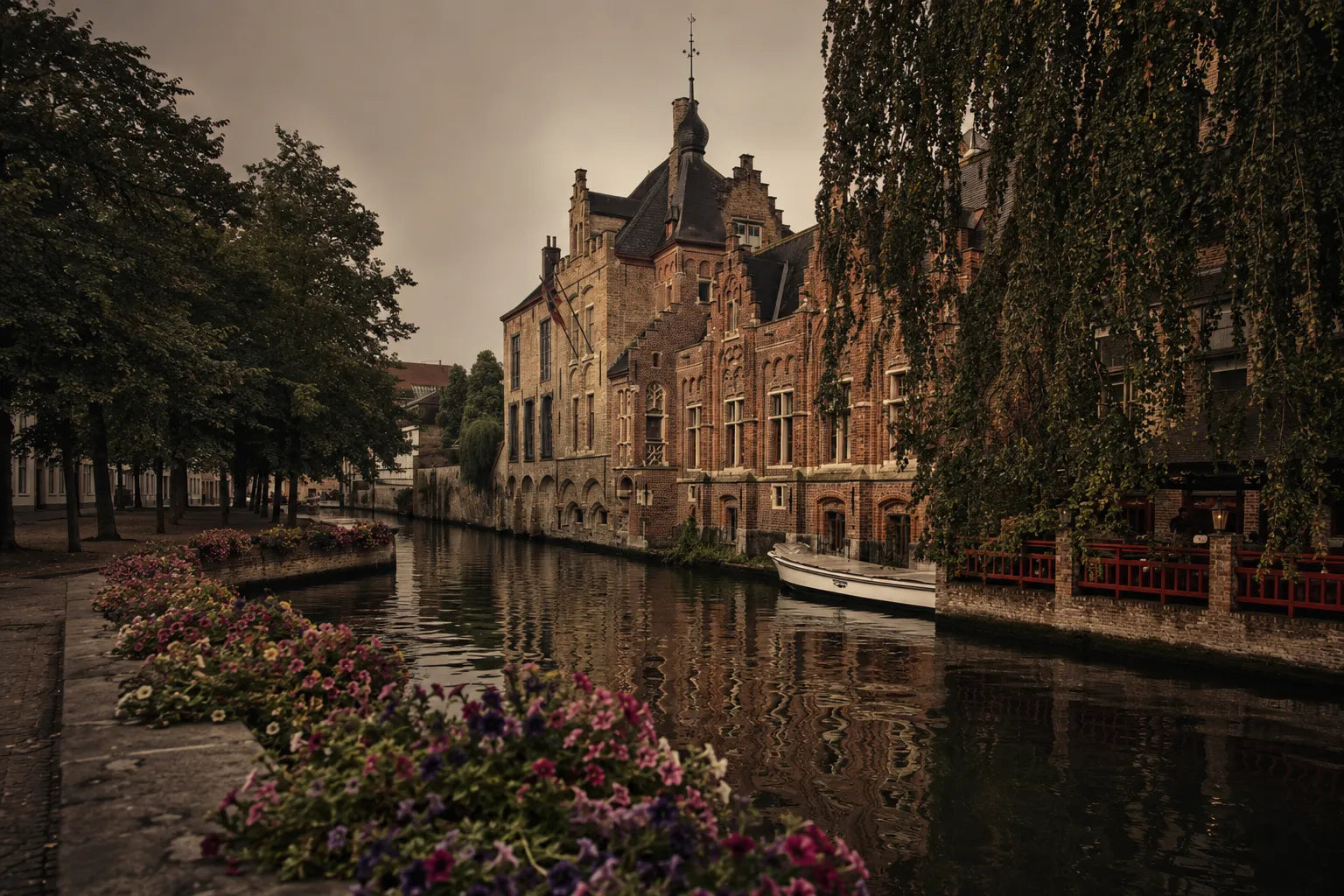 A moody, sepia-toned photograph of a canal lined with historic brick buildings, trees, and pink and purple flowers in the foreground.