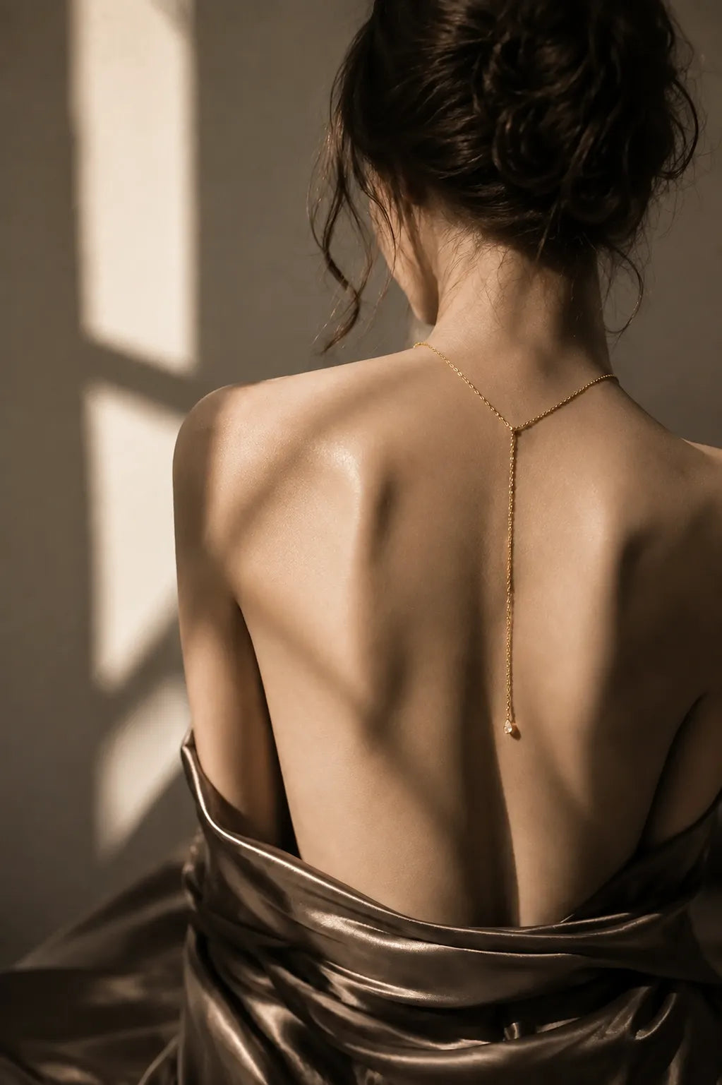 A woman shown from behind wearing a delicate gold Y-shaped chain necklace with a small pendant, draped in dark satin fabric against a dark background with dramatic window light shadows.