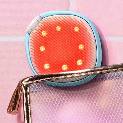 A small oval LED light therapy device with a light blue border and glowing orange-red LEDs, shown resting on a pink textured surface next to a metallic mesh pouch.