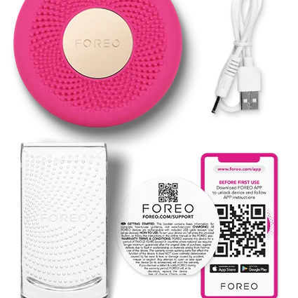 A FOREO handheld facial cleansing device in bright pink with a gold-toned center, shown with a white USB charging cable, a clear glass cup, and a printed quick-start card on a white background.