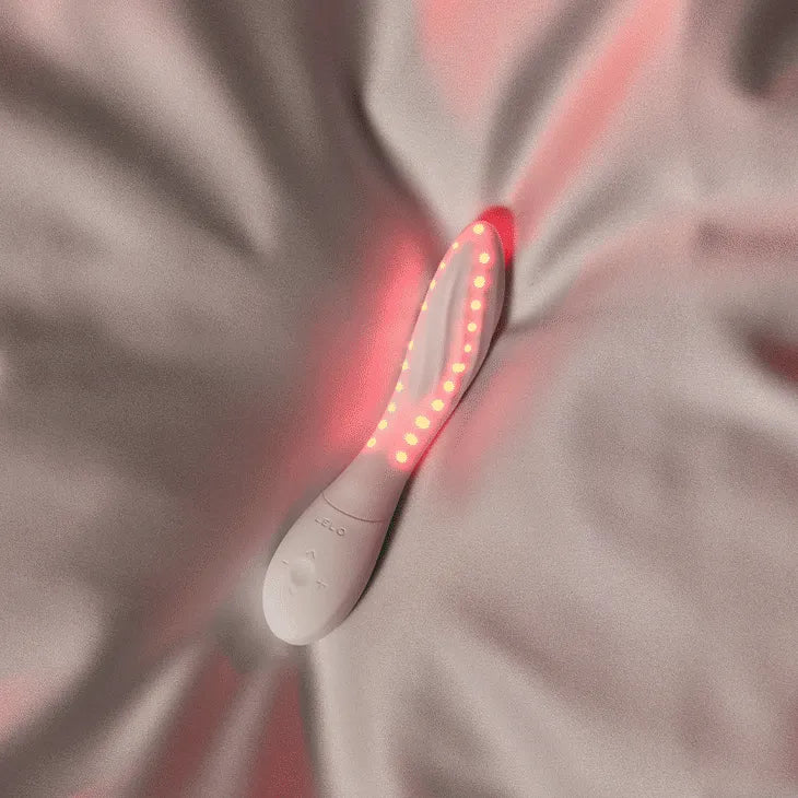 A white handheld LED light therapy device with glowing red lights, resting on a draped satin fabric background.