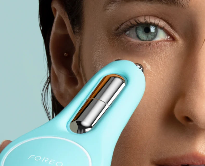 Close-up of a woman using a turquoise skincare device on her face against a light blue background.