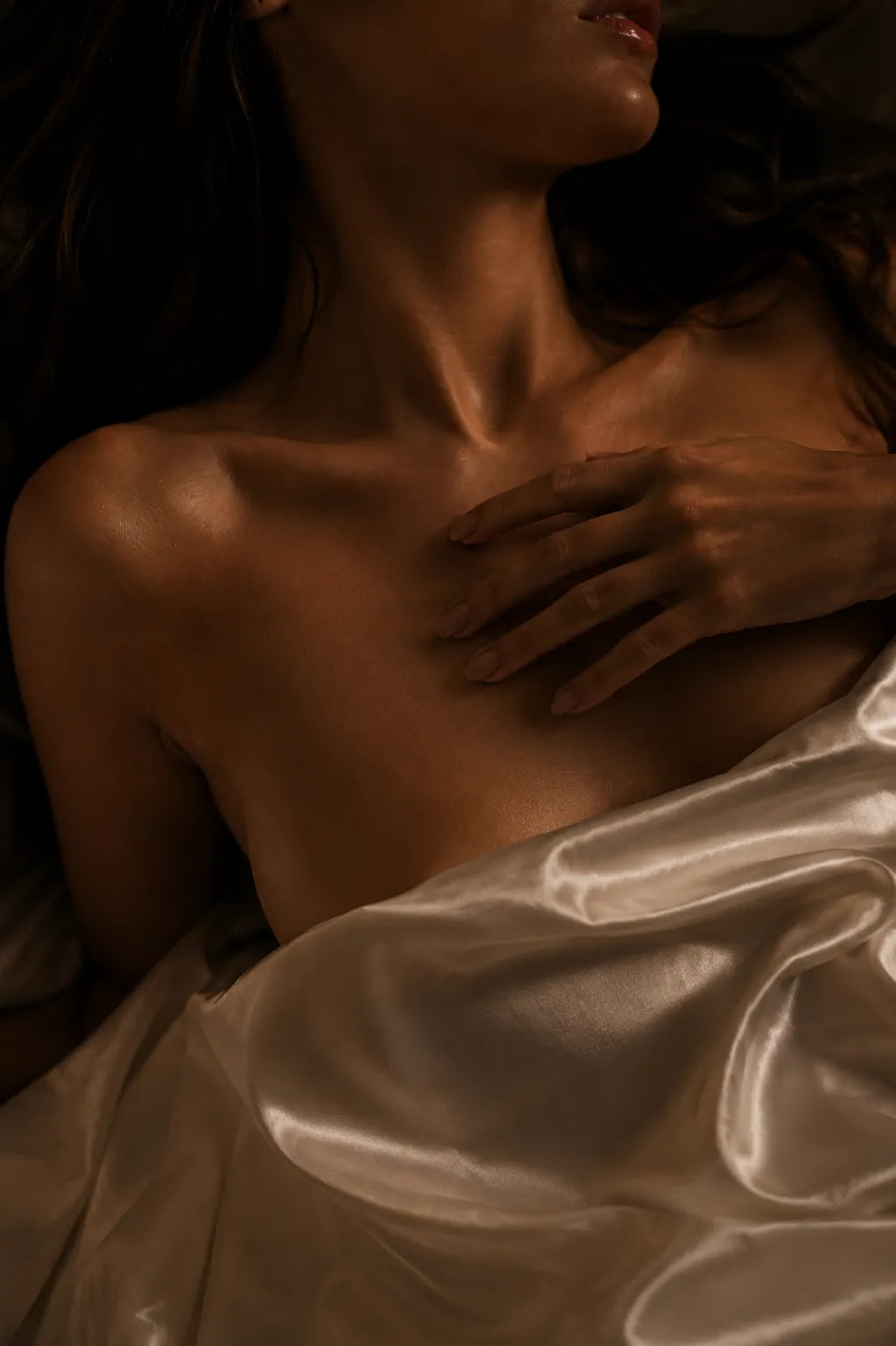 Close-up of a woman's bare chest and hand under warm, dim lighting, draped with shiny satin fabric.